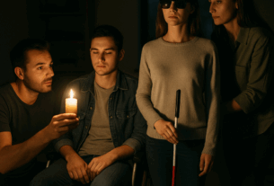 Vier Personen befinden sich in einem dunklen Raum während eines Stromausfalls. Links kniet ein junger Mann und hält eine Kerze in der Hand, deren Licht die Szene sanft beleuchtet. Daneben sitzt ein junger Mann im Rollstuhl mit ruhigem, nachdenklichem Blick. Rechts steht eine junge blinde Frau mit dunkler Sonnenbrille und einem weißen Langstock – sie steht aufrecht und ruhig. Hinter ihr steht eine weitere Frau, leicht nach vorn gebeugt, aufmerksam und bereit zu helfen. Der Hintergrund ist dunkel, die Stimmung ernst, aber ruhig. Das Bild vermittelt Fürsorge, Zusammenhalt und stille Wachsamkeit in einer Krisensituation.