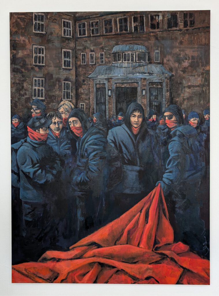 Eine Gruppe junger Menschen steht eng beieinander vor einem monumentalen, braunen Backsteingebäude. Sie tragen dunkle Kapuzenjacken, viele haben rote Tücher über Mund und Nase gezogen. Der Blick einiger richtet sich direkt nach vorn – ruhig, aber bestimmt. Im Vordergrund liegt ein ausgebreitetes, knallrotes Tuch oder Banner in Falten auf dem Boden. Die Szene wirkt angespannt, aber nicht gewalttätig – als würde etwas vorbereitet oder gleich beginnen. Die Architektur im Hintergrund erinnert an eine Schule oder ein Behördengebäude, was der Szene eine zusätzliche Schwere verleiht.